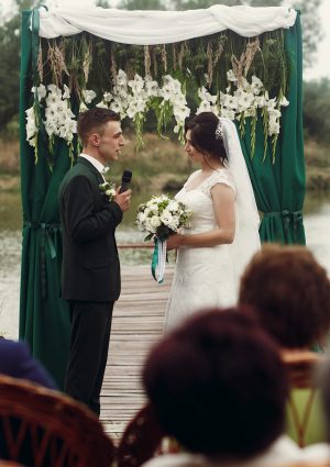 Handsome emotional groom in stylish suit giving wedding vow to beautiful bride with bouquet at outdoor wedding ceremony near aisle and lake, guests in the foreground